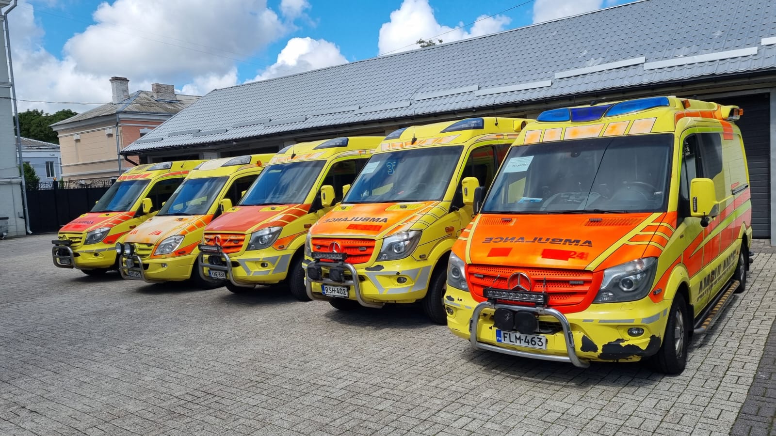 Rivi kelta-oransseja ambulansseja valmiina lähtöön asfalttipihalla.